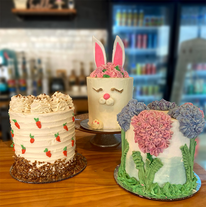 Three cakes on the counter, each easter themed. The cake on the left is a carrot cake with small carrots piped onto the sides in icing. The center cake is decorated to look like a bunny, with a cute face on the side and ears sticking out of the top. The third cake has hydrangeas piped in icing all over the cake.