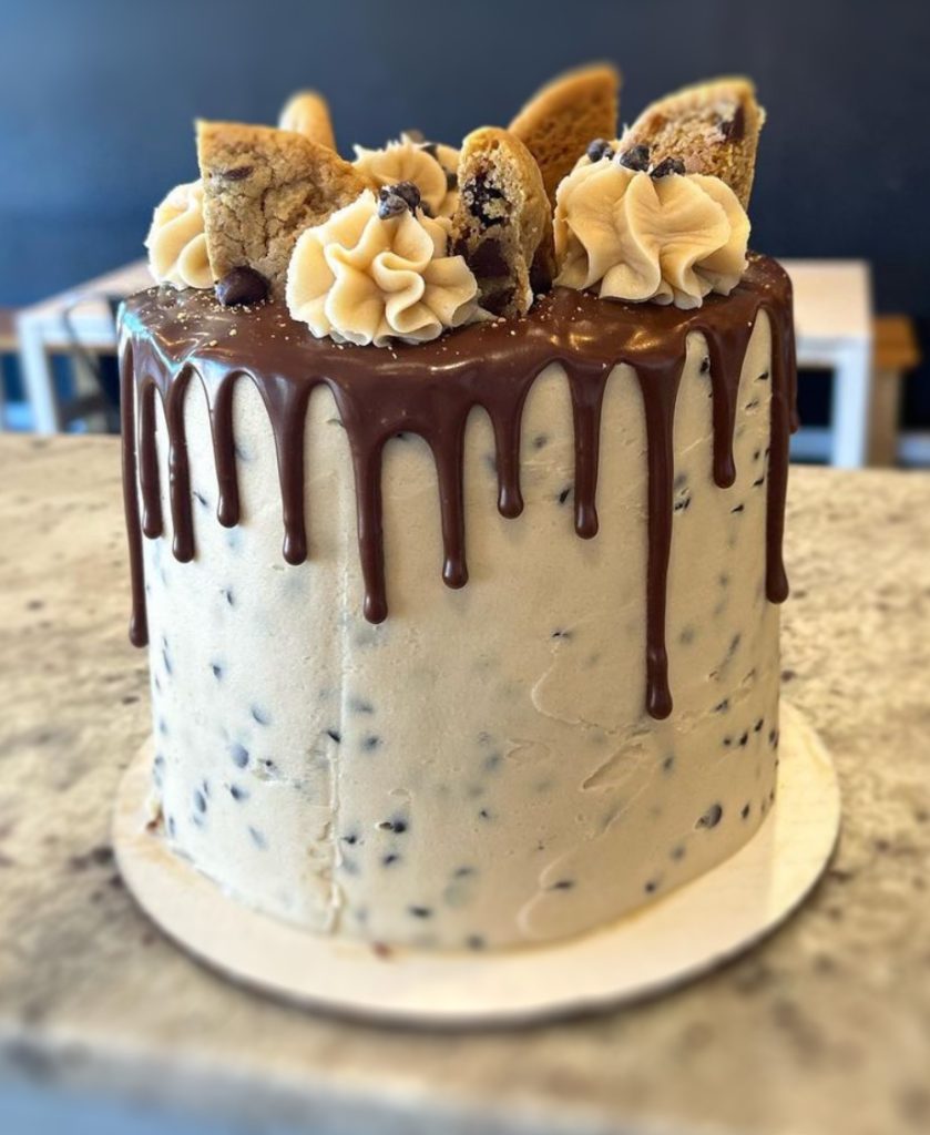Cake with Chocolate chip cookies on top and chocolate sauce dripping down the sides in a stylized way. Icing all over the cake has small chocolate chips in it that look like cookie dough.